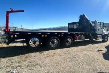 Flatbed work truck with knuckle boom crane mounted behind the cab, parked on gravel.