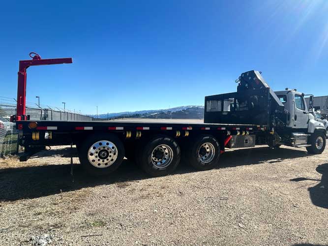 Flatbed work truck with knuckle boom crane mounted behind the cab, parked on gravel.