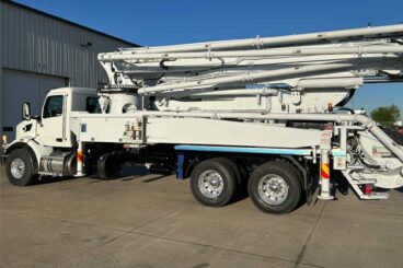 White concrete boom pump with dual rear axles parked outside industrial shop garage door