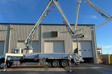 Industrial concrete boom pump truck parked outside shop doors. Boom shows ZR folding configuration with outriggers extended.