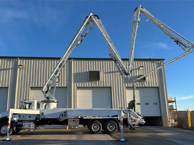 Industrial concrete boom pump truck parked outside shop doors. Boom shows ZR folding configuration with outriggers extended.