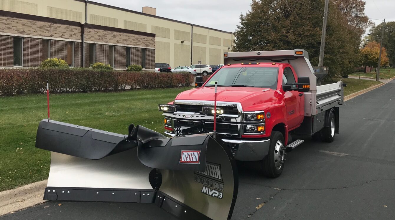 Front mounted Western MVP3 adjustable v-plow with dump body parked next to a curb outside an office building.