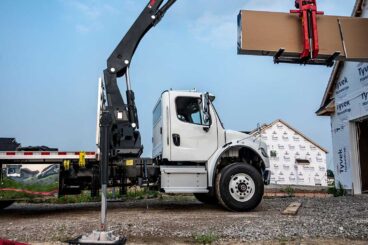 Truck with HIAB knuckle boom crane lifting and loading drywall materials into a window on a home construction job site.