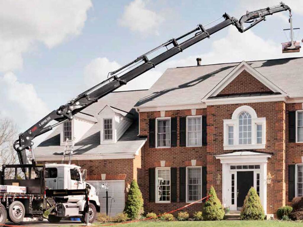 Flatbed work truck with HIAB heavy-duty knuckle boom crane parked in the driveway with extended boom over the roof of a home.