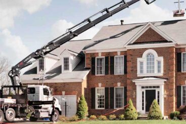 Flatbed work truck with HIAB heavy-duty knuckle boom crane parked in the driveway with extended boom over the roof of a home.