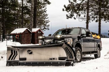 Pickup with front-mounted stainless steel FISHER wing plow and salt spreader in the truck bed parked on a snowy driveway. Wood cabins are visible in the background.