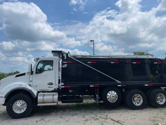 Large industrial dump truck with two rear axles and a pusher axle parked on concrete under cloudy skies.