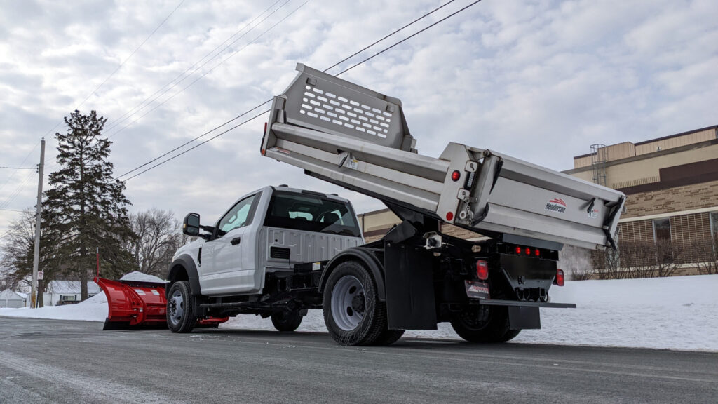 Truck with snow plow and raised dump bed parked on the street in front of a business.