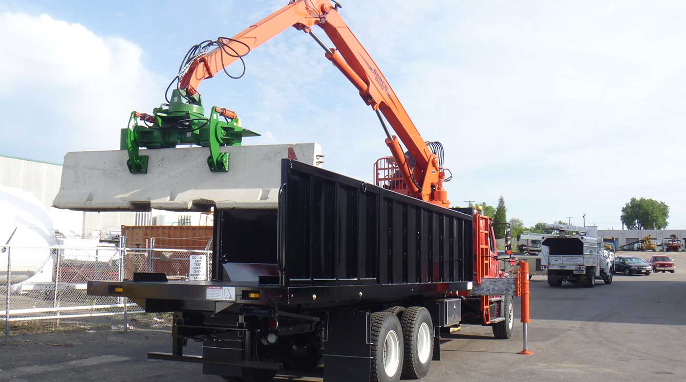 Industrial truck parked on asphalt lot. Orange loader with green grapple attachment lifting a concrete barrier from the truck body. Serco logo on the crane.